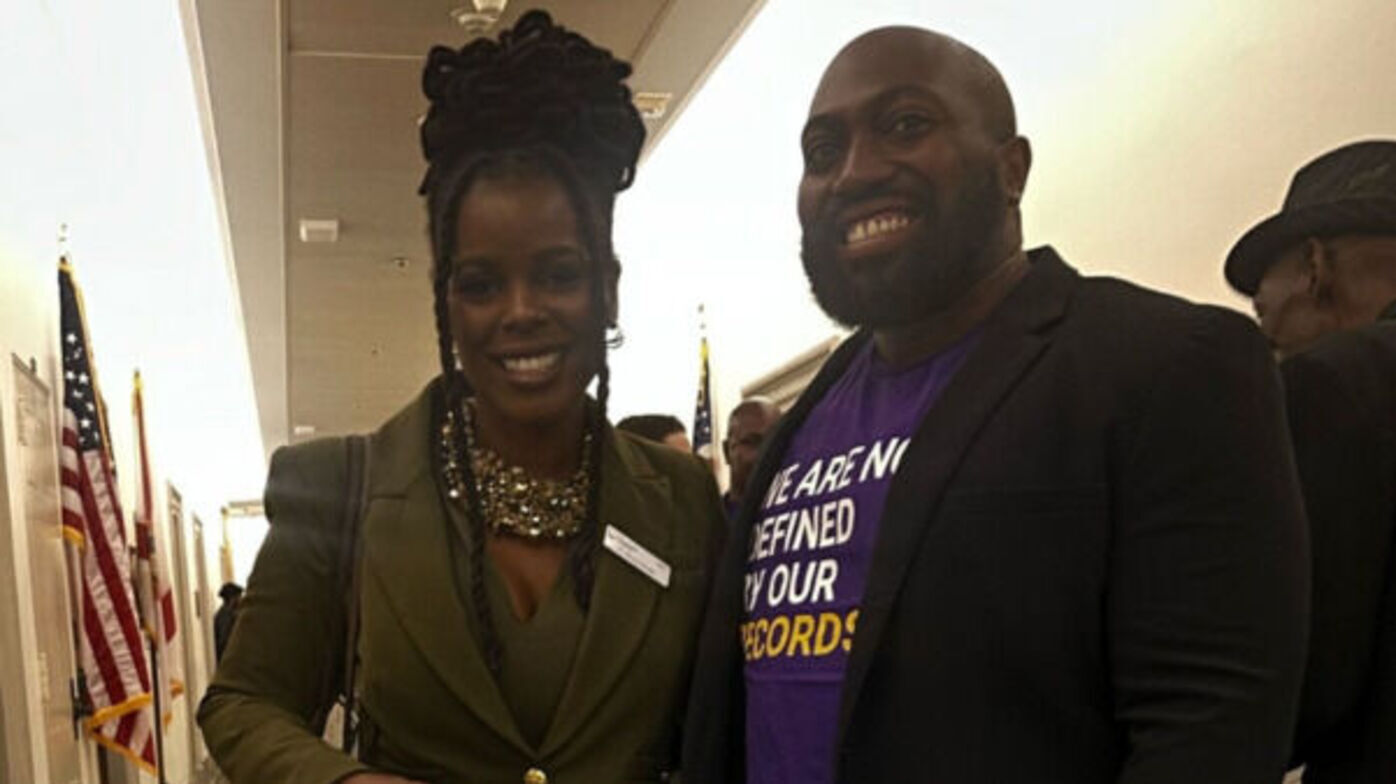 Two individuals are standing side by side in an indoor hallway. The woman on the left is wearing a fitted olive green suit and a statement necklace, smiling and holding a patterned handbag. The man on the right is also smiling and wearing a dark blazer over a purple t-shirt with the message 
